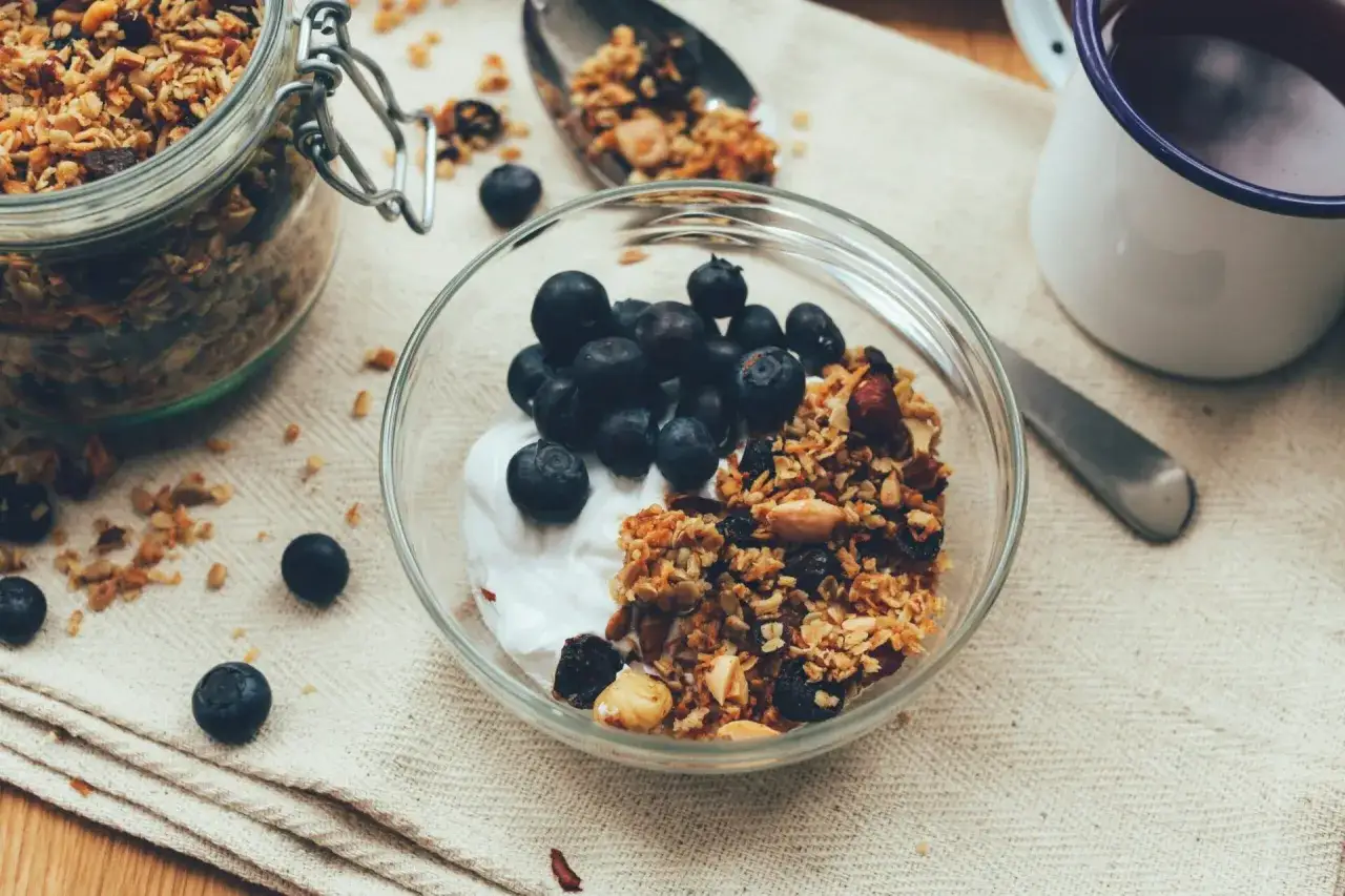 En sk&aring;l med kr&auml;mig smaksatt kvarg, toppad med bl&aring;b&auml;r och granola. En burk med granola och en mugg kaffe st&aring;r bredvid.