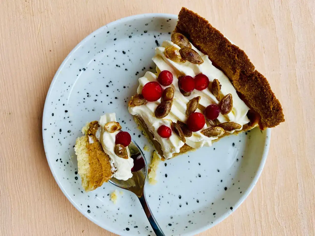 En bit glutenfri pumpapaj med vispgr&auml;dde, lingon och rostade pumpafr&ouml;n p&aring; en prickig tallrik. En sked med en bit paj ligger bredvid.