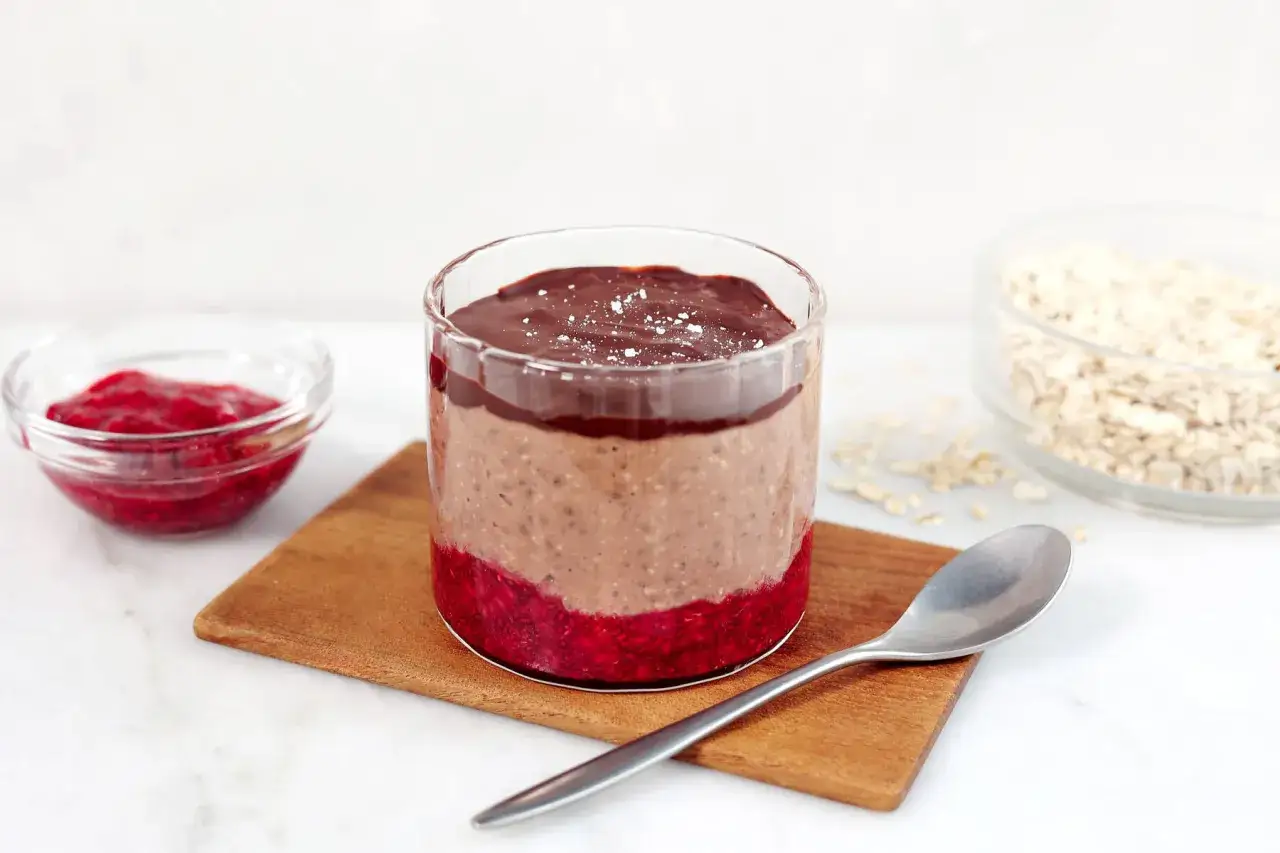 En glasburk med overnight oats med chokladsmak, hallonsylt och en sked. Bredvid finns en sk&aring;l med hallon och en sk&aring;l med havregryn.